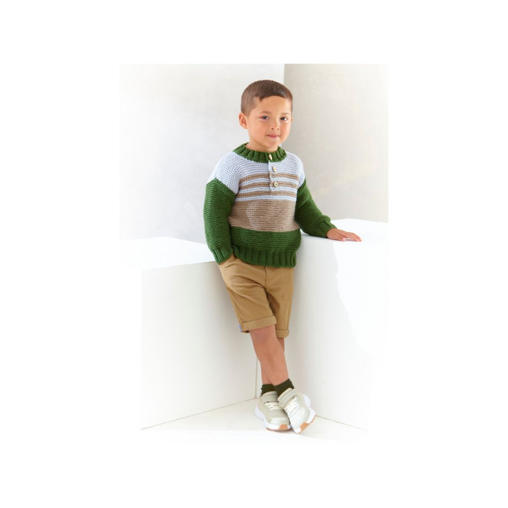 A young boy wearing a green, beige, and white striped knit sweater, khaki shorts, and white sneakers, leaning against a white architectural corner.