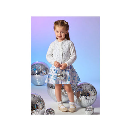 A young girl stands in a studio wearing a sparkly white long-sleeve cardigan and a light blue patterned skirt, surrounded by several large mirrored disco balls against a soft blue-to-purple gradient background.