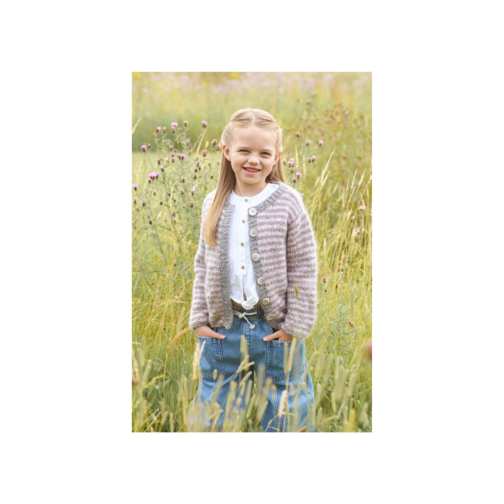 A young girl with blonde hair stands in a field of tall grass, wearing a textured grey and white striped cardigan over a white top and blue jeans.