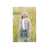 A young girl with blonde hair stands in a field of tall grass, wearing a textured grey and white striped cardigan over a white top and blue jeans.