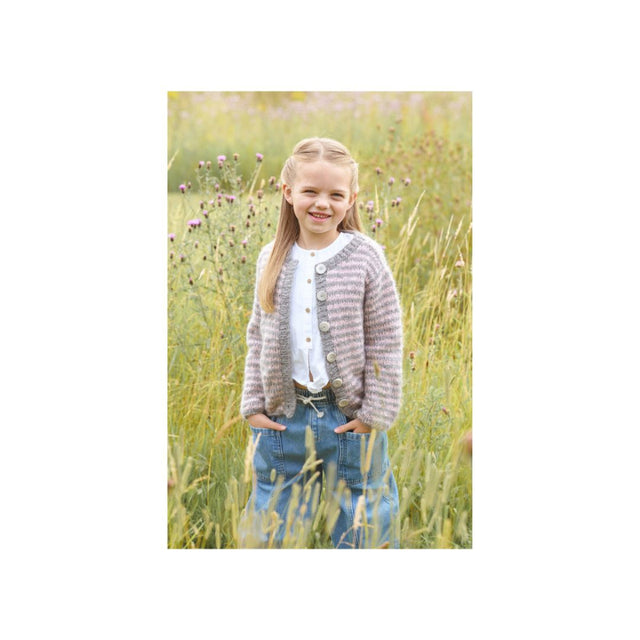A young girl with blonde hair stands in a field of tall grass, wearing a textured grey and white striped cardigan over a white top and blue jeans.