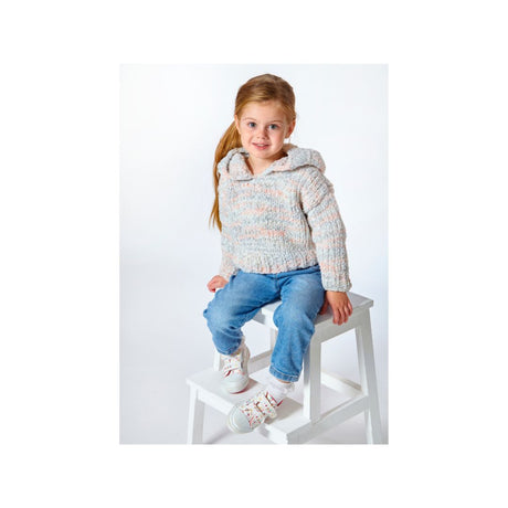 A young girl with blonde hair in a ponytail sits on a white wooden stool against a plain white studio background. She is wearing a chunky, textured knit hooded sweater in a heathered light gray, pink, and blue colorway, paired with blue jeans and white sneakers.