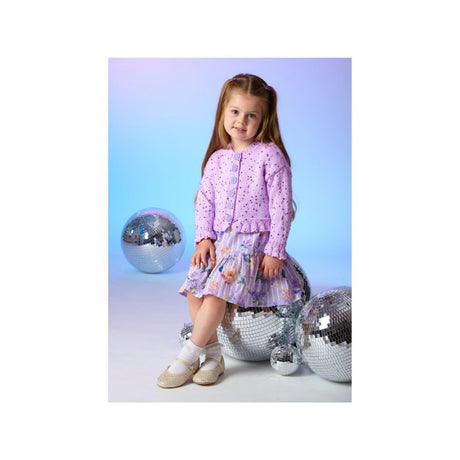 A young girl with long light brown hair sits on a silver disco ball, wearing a light purple knit cardigan and a matching purple patterned skirt. She is posed with two large silver disco balls against a blue and purple gradient studio background.