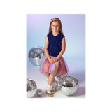 A young girl with a pink hair bow sits on a white floor surrounded by large silver disco balls, wearing a sleeveless navy blue sequined top and a pink and purple tulle skirt.