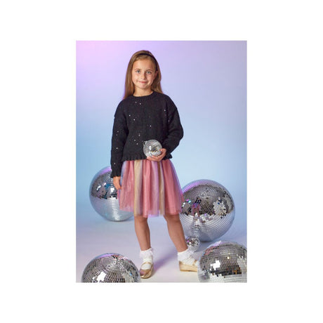 A young girl stands in a studio wearing a black sequined long-sleeve top and a pink tulle skirt, holding a silver disco ball. Several large mirrored disco balls are placed around her on a pastel blue-to-purple gradient background.