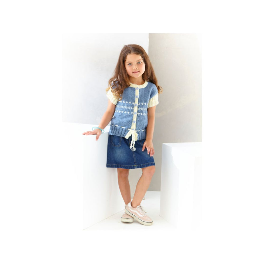 A young girl with long brown hair stands in a white architectural setting, wearing a light blue and white patterned knit vest over a cream t-shirt, a blue denim skirt, and beige platform sneakers.