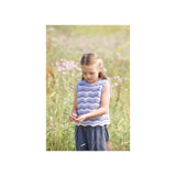 A young girl standing in a sunlit field of wildflowers, wearing a sleeveless lavender and white patterned knit top with a scalloped hem and a dark skirt.