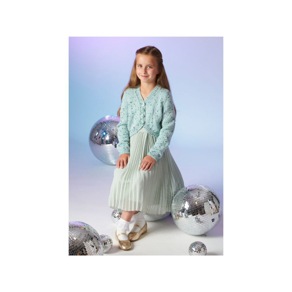 A young girl stands in a studio wearing a light mint green knit cardigan over a matching pleated skirt. She is posed among several large silver disco balls against a soft blue-to-lavender gradient background.