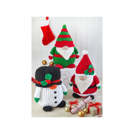 Three knitted Christmas gnome and snowman decorations displayed on a tabletop with a red stocking, ornaments, wrapped gifts, and a white paneled backdrop.