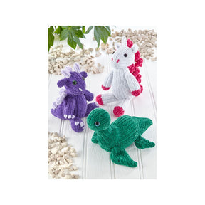 Three knitted plush fantasy creatures on a white wood surface: a purple dragon, a white unicorn with pink details, and a green loch ness monster. Small beige pebbles and green leaves are arranged around them.