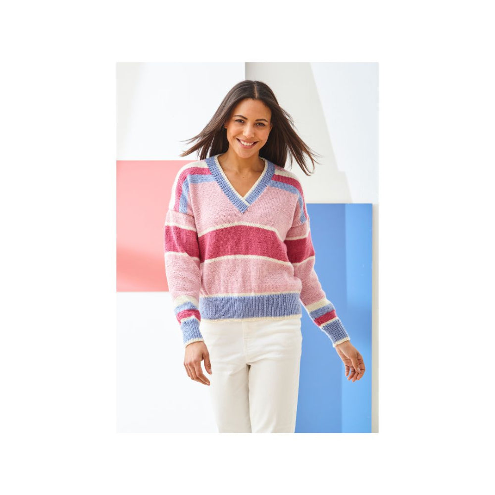 A woman wearing a pink, red, and blue striped knit sweater with a V-neck and white pants, standing against a modern geometric background of white, pink, and blue panels.