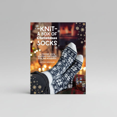 A book or kit cover titled "Knit- A Box of Christmas Socks" showing a pair of knitted Christmas socks on feet against a warm holiday background with text and snowflake graphics.