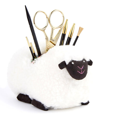 A white sheep-shaped holder with a black face and black hooves, filled with black and gold knitting accessories, on a white background.