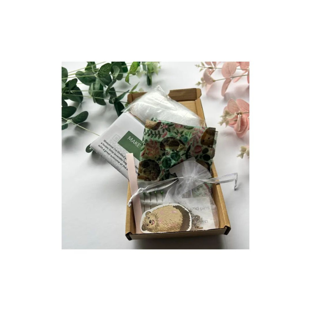 A small cardboard gift box on a white surface containing a patterned fabric pouch, a clear plastic bag, and printed paper inserts. Green leaves and pale pink flowers are placed around the box as styling props.
