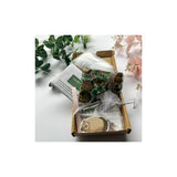 A small cardboard gift box on a white surface containing a patterned fabric pouch, a clear plastic bag, and printed paper inserts. Green leaves and pale pink flowers are placed around the box as styling props.