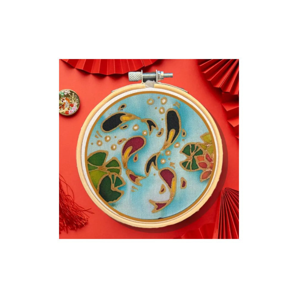 A circular embroidery hoop holding light blue fabric with a colourful floral and koi fish design, surrounded by red decorative paper fans and tassels on a white background.