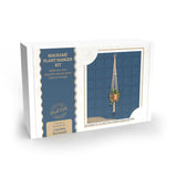A boxed macramé plant hanger kit shown at a slight angle on a white background. The package is white with a navy blue panel and a photo of a hanging macramé plant holder.