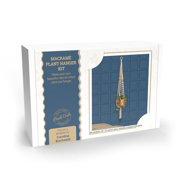 A boxed macramé plant hanger kit shown at a slight angle on a white background. The package is white with a navy blue panel and a photo of a hanging macramé plant holder.