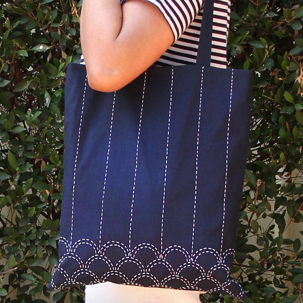 A navy blue canvas tote bag with white vertical dashed stripes and a scalloped hem, carried on the shoulder of a person wearing a navy and white striped shirt, set against a background of green foliage.