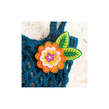 A colourful embroidered flower and leaf applique brooch pinned onto a dark teal crocheted bag, set against a light beige textured background.