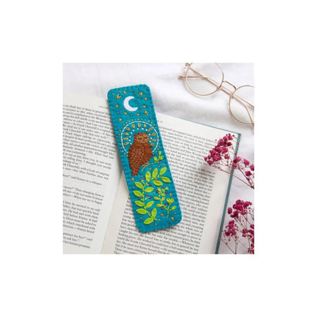 A turquoise felt bookmark with a brown owl, green leaves, and a crescent moon design, resting on an open book with eyeglasses and pink flowers as props.
