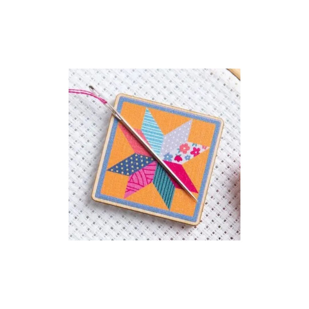 A square cross-stitch sampler featuring a colorful geometric star design on a white mesh fabric, with a wooden needle loom visible at the top and a silver needle resting diagonally across it.