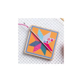 A square cross-stitch sampler featuring a colorful geometric star design on a white mesh fabric, with a wooden needle loom visible at the top and a silver needle resting diagonally across it.