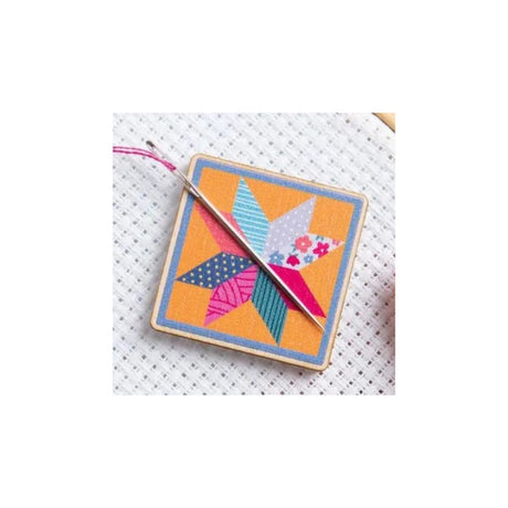 A square cross-stitch sampler featuring a colorful geometric star design on a white mesh fabric, with a wooden needle loom visible at the top and a silver needle resting diagonally across it.