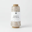 A spool of Rico Design Creative Make It Pailleçte yarn thread, featuring a white paper label with black text and silver sequins on a light beige thread wound around a brown cardboard core, set against a plain white background.