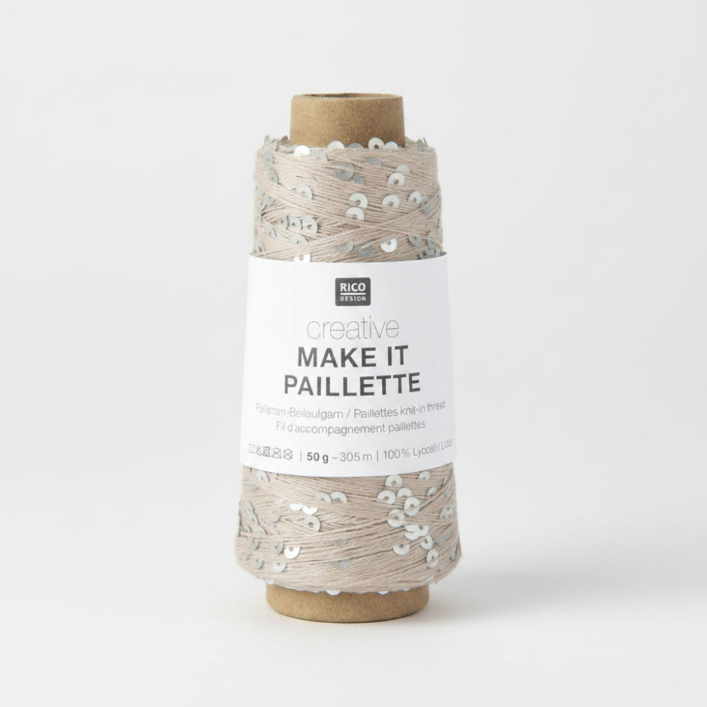 A spool of Rico Design Creative Make It Pailleçte yarn thread, featuring a white paper label with black text and silver sequins on a light beige thread wound around a brown cardboard core, set against a plain white background.