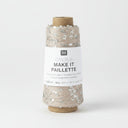 A spool of Rico Design Creative Make It Pailleçte yarn thread, featuring a white paper label with black text and silver sequins on a light beige thread wound around a brown cardboard core, set against a plain white background.