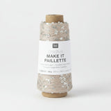 A spool of Rico Design Creative Make It Pailleçte yarn thread, featuring a white paper label with black text and silver sequins on a light beige thread wound around a brown cardboard core, set against a plain white background.