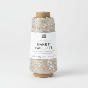 A spool of Rico Design Creative Make It Pailleçte yarn thread, featuring a white paper label with black text and silver sequins on a light beige thread wound around a brown cardboard core, set against a plain white background.