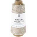 A spool of Rico Design Creative Make It Pailleçte yarn thread, featuring a white paper label with black text and silver sequins on a light beige thread wound around a brown cardboard core, set against a plain white background.