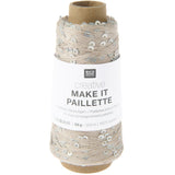 A spool of Rico Design Creative Make It Pailleçte yarn thread, featuring a white paper label with black text and silver sequins on a light beige thread wound around a brown cardboard core, set against a plain white background.
