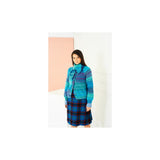 A woman modeling a colourful striped knit cardigan and matching scarf in shades of blue, teal, and purple, paired with a blue and red plaid skirt, set against a minimalist white and pastel pink background.