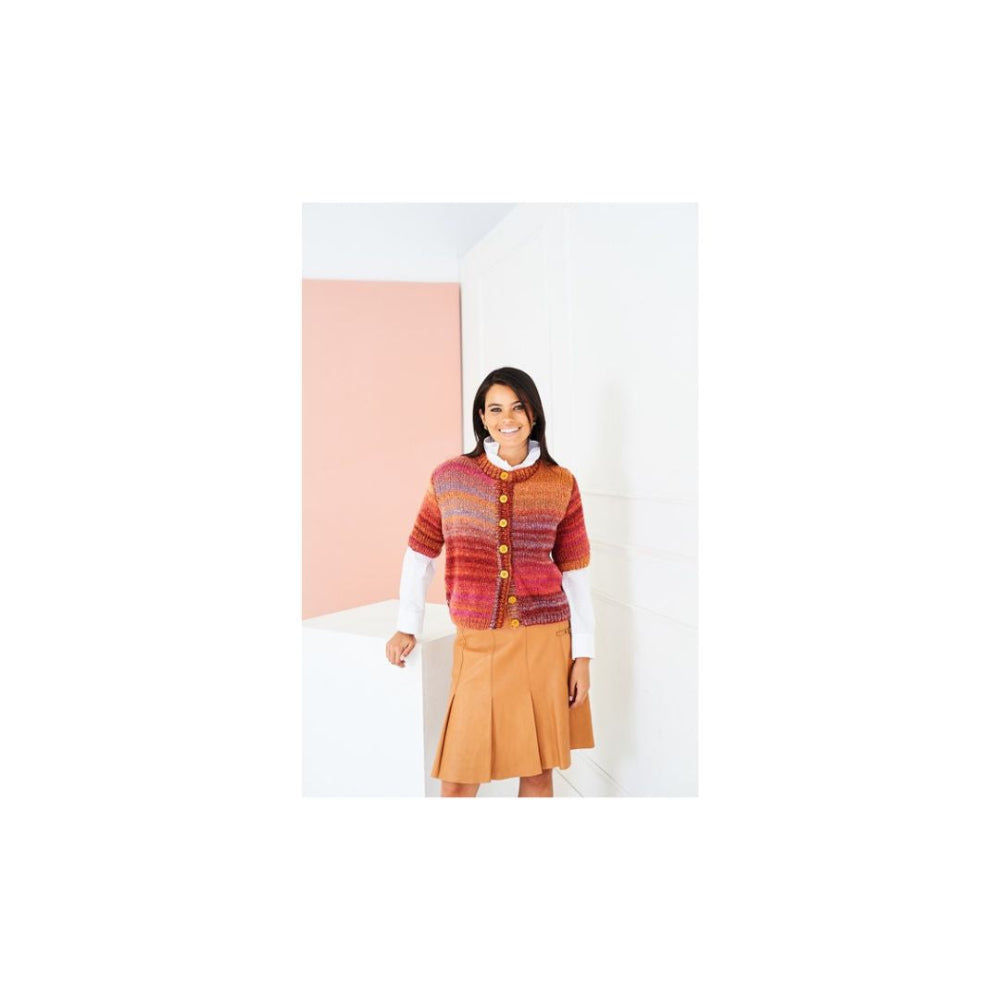 A woman modeling a colourful striped knit cardigan with buttons over a white long-sleeve shirt and a tan pleated skirt, standing in a minimalist studio setting with white walls and a soft pink accent panel.