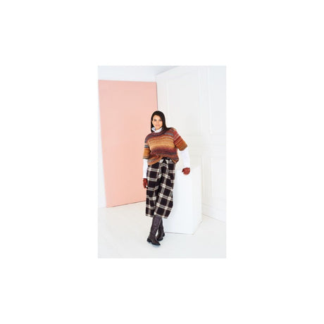 A model stands in a minimalist studio setting wearing a striped knit sweater, a plaid skirt, and dark boots, with a large pink panel and white walls in the background.