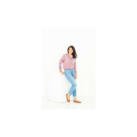 A woman modeling a colourful patterned crochet cardigan and light blue jeans, leaning against a white block on a white studio background.