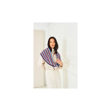 A woman wearing a cream-coloured outfit and a striped crochet wrap shawl in red, blue, and white, standing in a minimalist white room.
