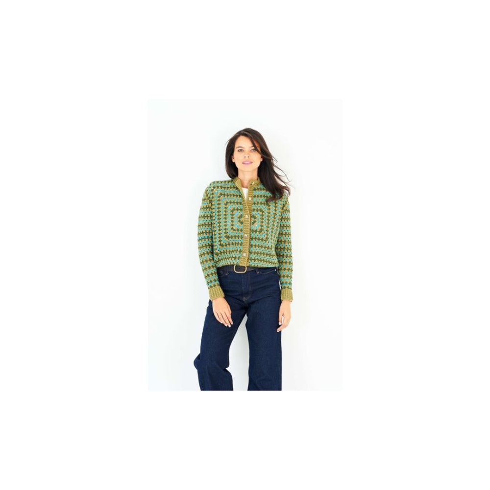 A woman modeling a green and yellow geometric patterned crochet cardigan with dark blue jeans against a plain white background.