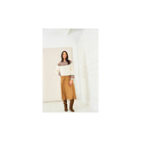 A woman stands in a bright white room wearing a cream crochet sweater with a multicolour striped yoke and cuffs, a tan skirt, and brown boots.