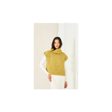 A woman wearing a textured mustard yellow crochet mock-neck sweater vest over a white long-sleeve top and white pants, standing against a minimalist off-white background.