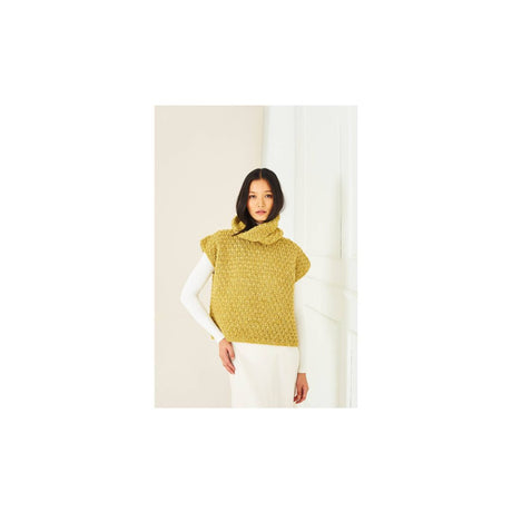 A woman wearing a textured mustard yellow crochet mock-neck sweater vest over a white long-sleeve top and white pants, standing against a minimalist off-white background.