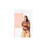 A woman wearing a multicolour knit sweater and a pleated beige skirt, standing against a white wall with a pink panel in the background.