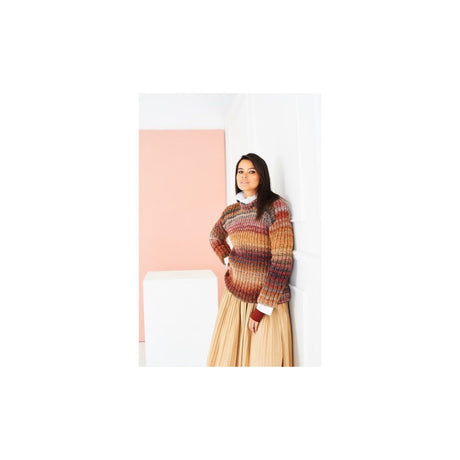 A woman wearing a multicolour knit sweater and a pleated beige skirt, standing against a white wall with a pink panel in the background.
