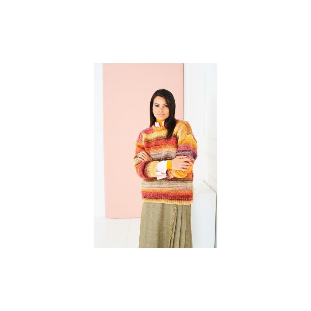 A woman stands with arms crossed wearing a multicoloured striped knit sweater in shades of orange, yellow, red, and brown, paired with a muted olive skirt, set against a minimalist pink and white background.