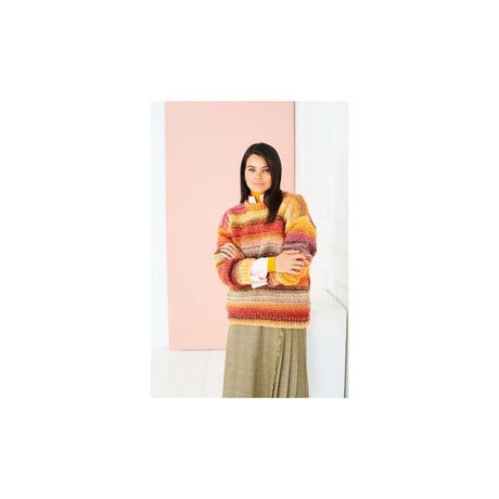 A woman stands with arms crossed wearing a multicoloured striped knit sweater in shades of orange, yellow, red, and brown, paired with a muted olive skirt, set against a minimalist pink and white background.