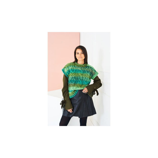 A woman modeling a green and blue patterned knit tank top over an olive green long-sleeve top and a dark skirt, set against a minimalist pink and white background.