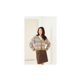 A woman modeling a colourful patterned knit cardigan with yellow trim over a white top and a brown skirt, standing against a plain off-white wall.